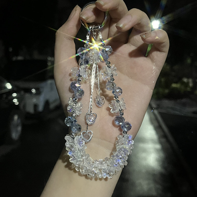 A hand holding a silver keychain with clear and light blue faceted crystal beads, heart charms, and a floral tassel against a dark nighttime outdoor background.